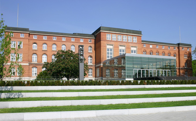 Backsteingebäude mit mehreren großen Fenstern und einem modernen Glasvorbau. Im Vordergrund sind Freitreppen und Grünflächen zu sehen. Der Himmel ist blau.