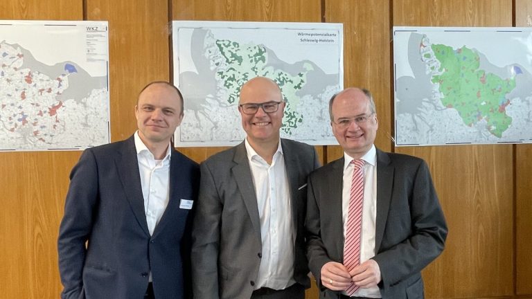 Drei Männer in Anzügen stehen in einem Konferenzraum vor holzverkleideten Wänden. An der Wand hängen Karten. Im Vordergrund sind schwarze Bürostühle und ein Tisch mit Dokumenten sichtbar. Ein Projektor ist an der Decke montiert.