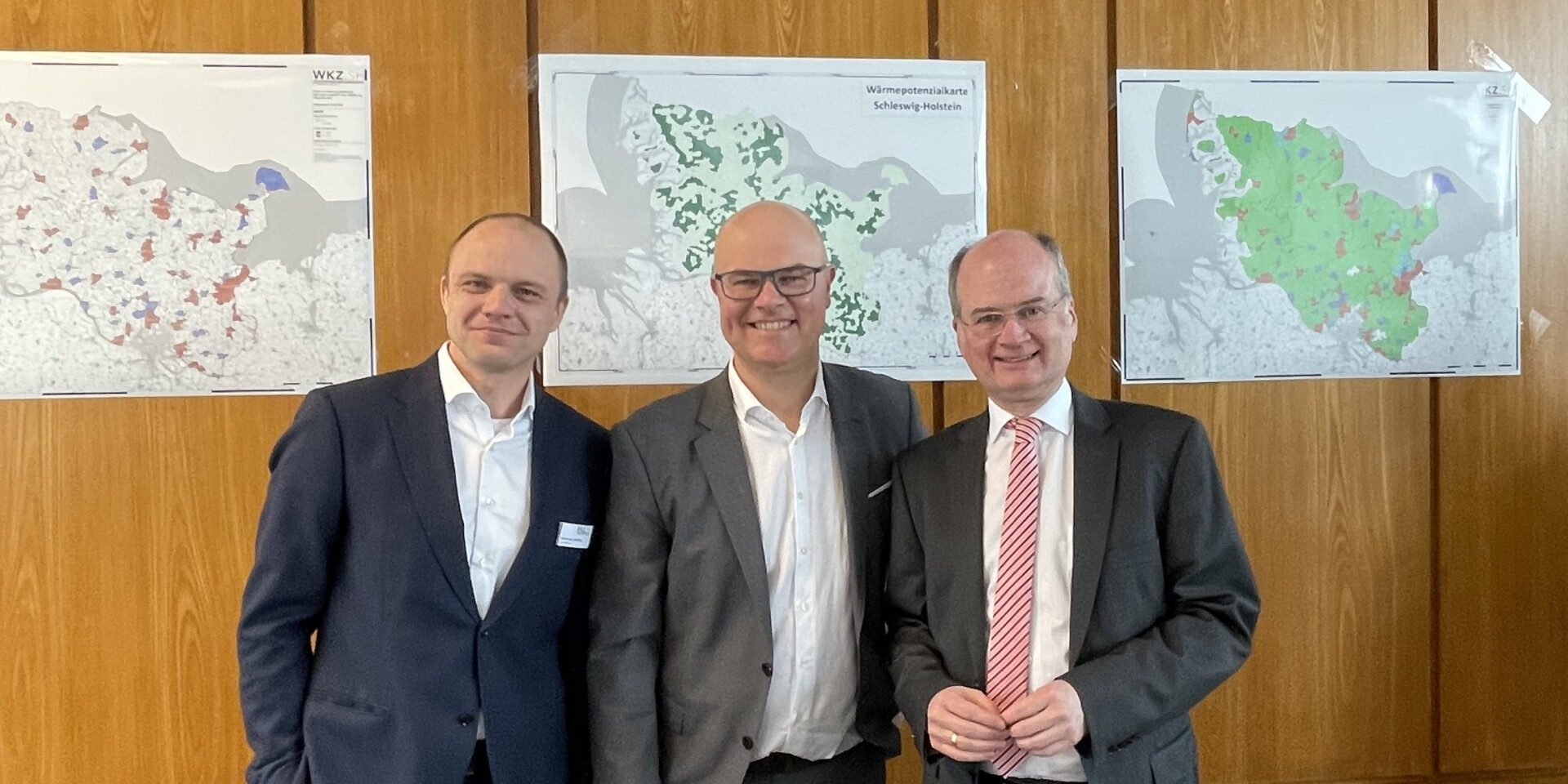 Drei Männer in Anzügen stehen in einem Konferenzraum vor holzverkleideten Wänden. An der Wand hängen Karten. Im Vordergrund sind schwarze Bürostühle und ein Tisch mit Dokumenten sichtbar. Ein Projektor ist an der Decke montiert.