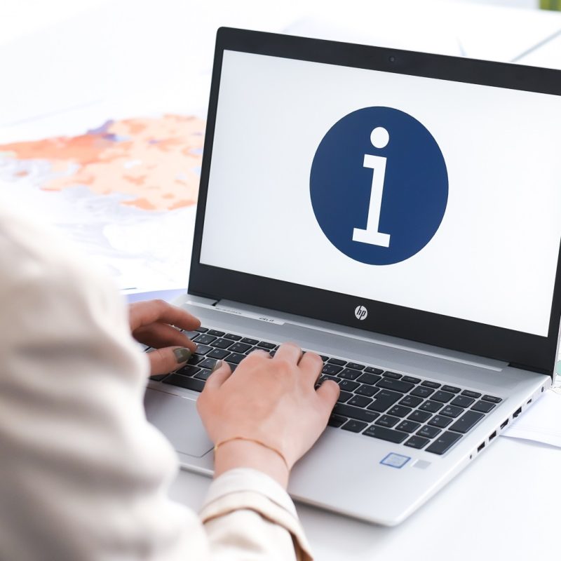Eine Person tippt auf einem Laptop mit einem auf dem Bildschirm angezeigten „i“-Symbol in einem blauen Kreis, welches auf Informationen hinweist. Der Hintergrund wirkt hell und unauffällig.
