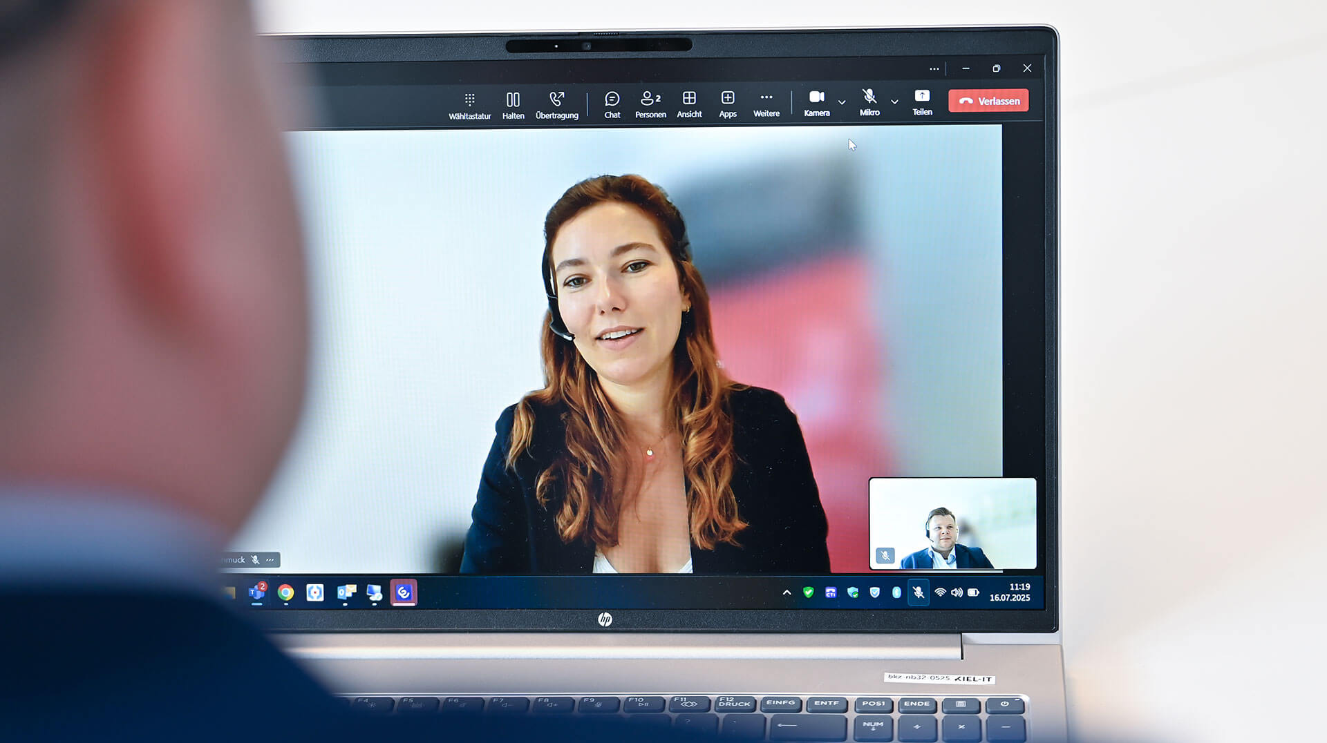 Ein Mann sitzt vor einem Laptop und führt ein virtuelles Meeting. Auf dem Bildschirm ist eine Frau sichtbar, die ein Headset trägt. Unten rechts gibt es ein kleines Vorschaufenster des Mannes.